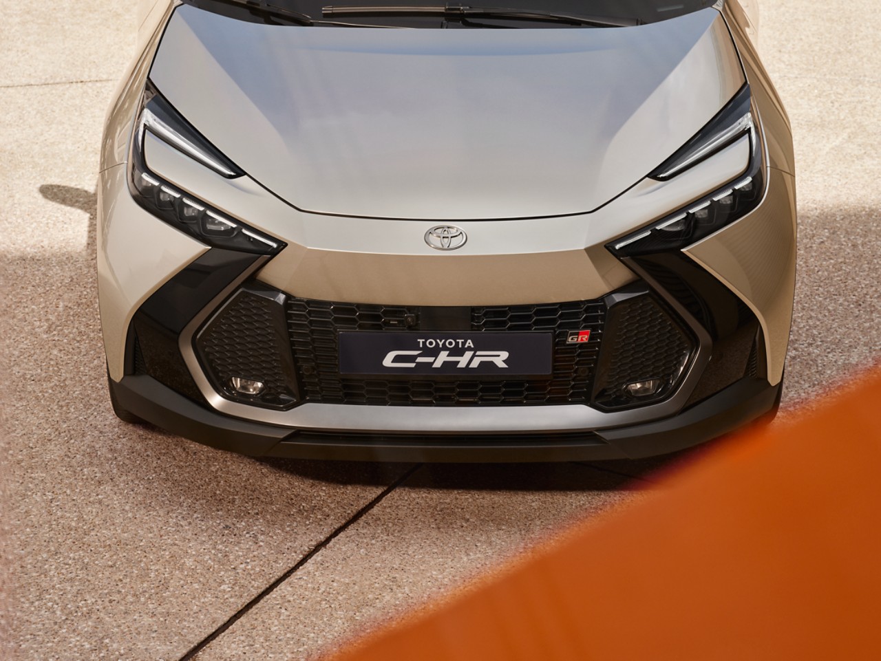 The front view of a New Toyota C-HR in silver with a black grille. It's parked on stone paving with a flash of orange in the foreground.